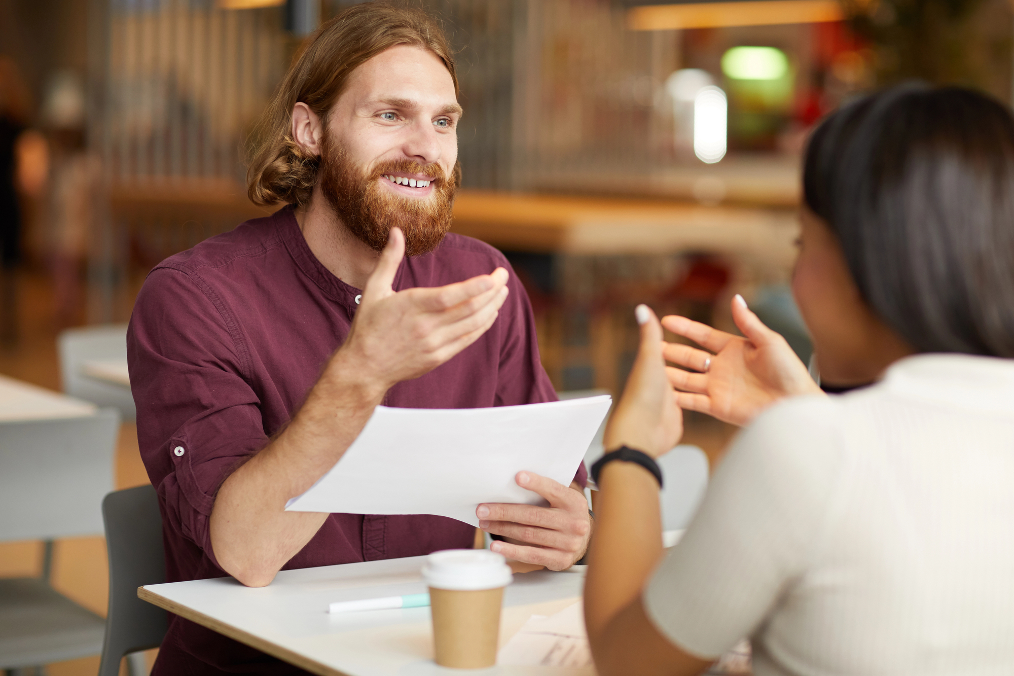 A bearded man in a maroon shirt is smiling and gesturing while holding papers during a conversation with another person, whose back is to the camera. They are sitting at a white table, with a coffee cup in the foreground.