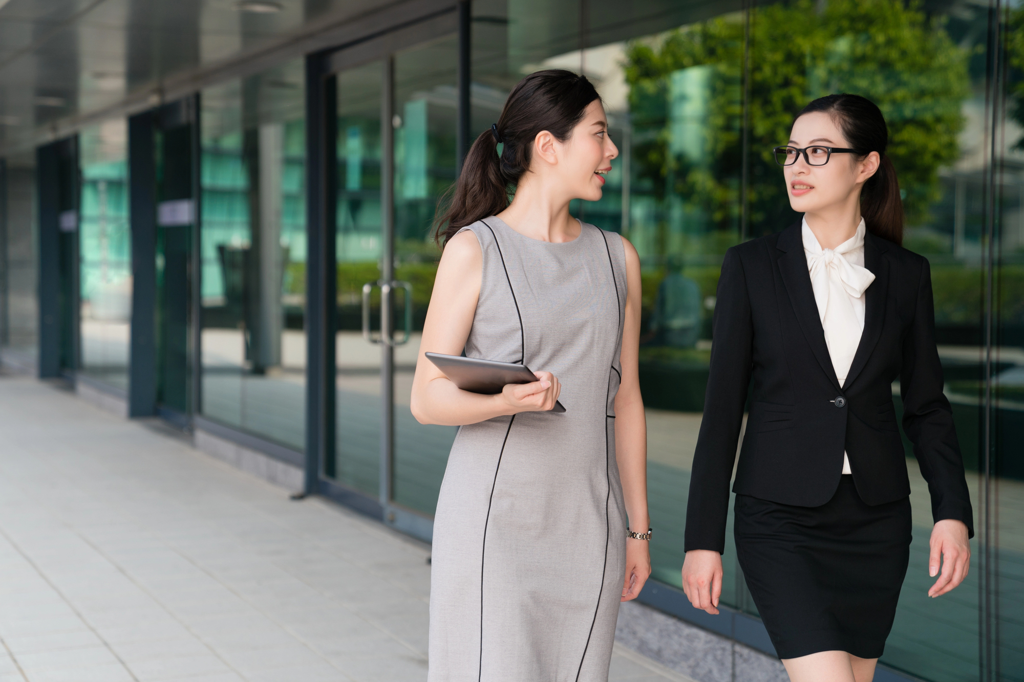 Two women walking outside a modern glass building. One wears a gray dress and holds a tablet, while the other wears a black suit with a white blouse. They are conversing and appear businesslike.