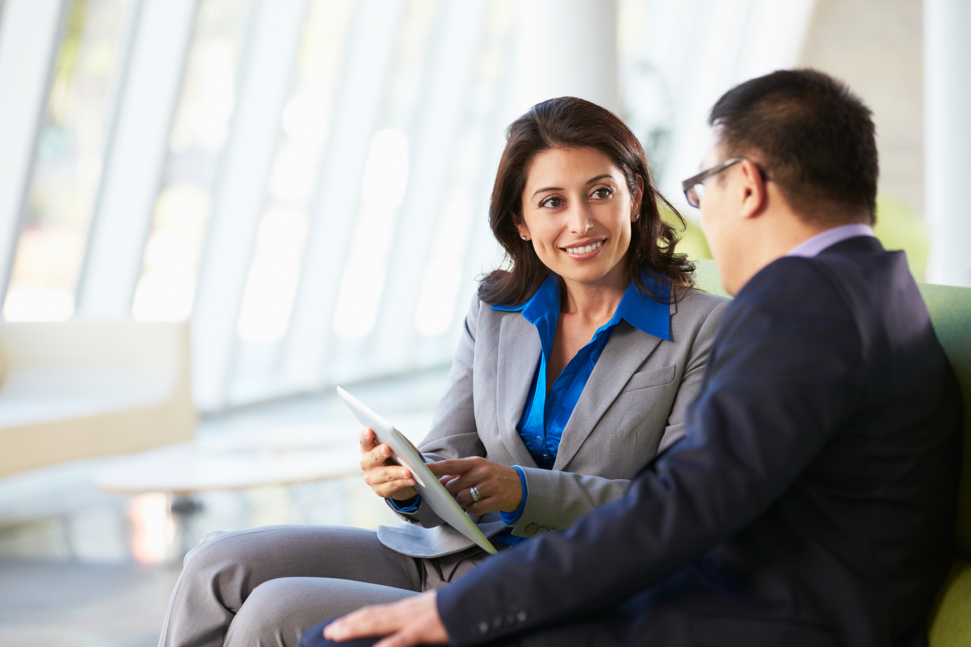 A woman and a man in business attire have a conversation while seated in a bright, modern office. The woman holds a tablet and smiles, engaging with the man, who wears glasses and faces her. Large windows provide natural light in the background.