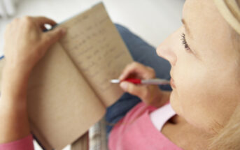 A person with long blonde hair, wearing a pink top, is writing in an open notebook with a red pen. The shot is taken from above and to the side, focusing on the writing hand and the notebook's pages.