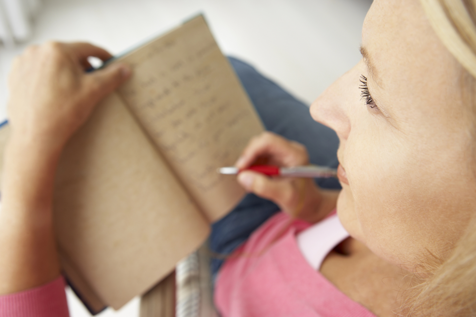 A person with long blonde hair, wearing a pink top, is writing in an open notebook with a red pen. The shot is taken from above and to the side, focusing on the writing hand and the notebook's pages.