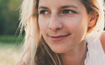 A woman with long blonde hair and a gentle smile looks off to the side. She is outdoors in a natural setting, with a blurred background of greenery, wearing a sleeveless white top.