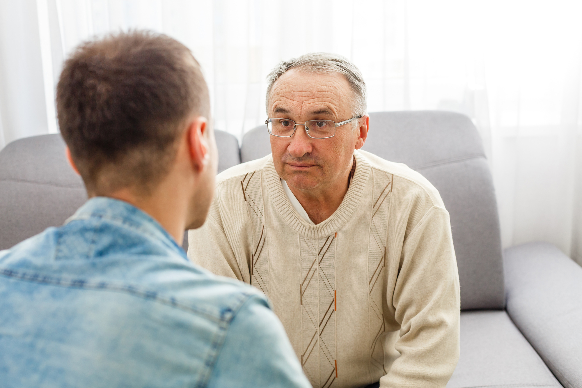 An older man wearing glasses and a light sweater is sitting on a gray couch, attentively talking to a younger man in a denim shirt. They are in a well-lit room with sheer curtains in the background.