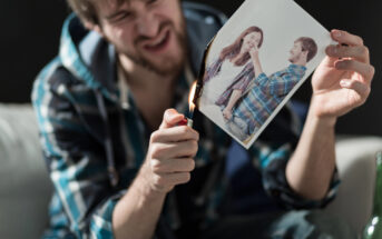 A man with a beard is sitting on a sofa, angrily burning a photo with a lighter. The photo shows a smiling couple. The background is dark and slightly out of focus, emphasizing the man's expression and actions.