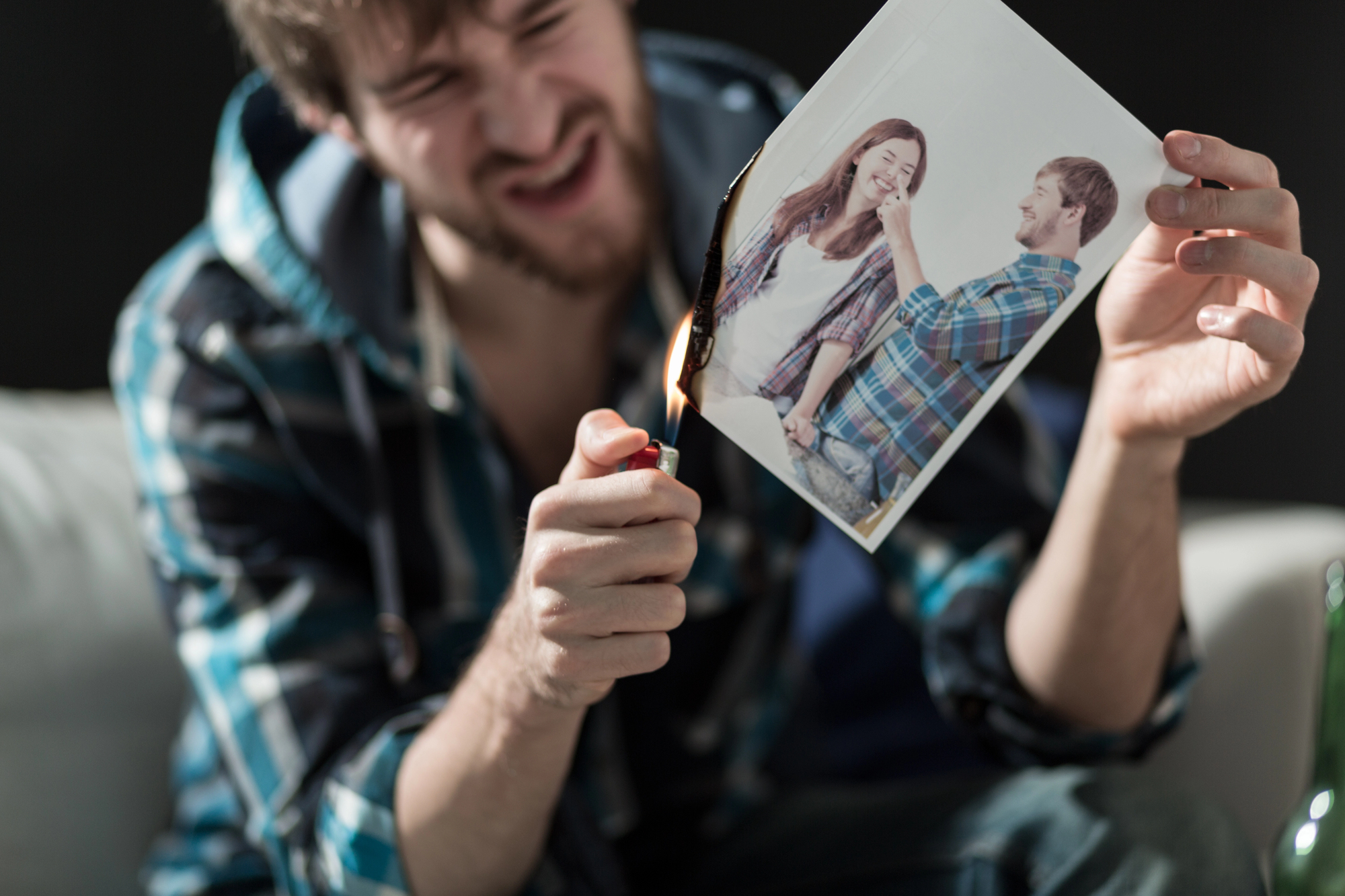 A man with a beard is sitting on a sofa, angrily burning a photo with a lighter. The photo shows a smiling couple. The background is dark and slightly out of focus, emphasizing the man's expression and actions.