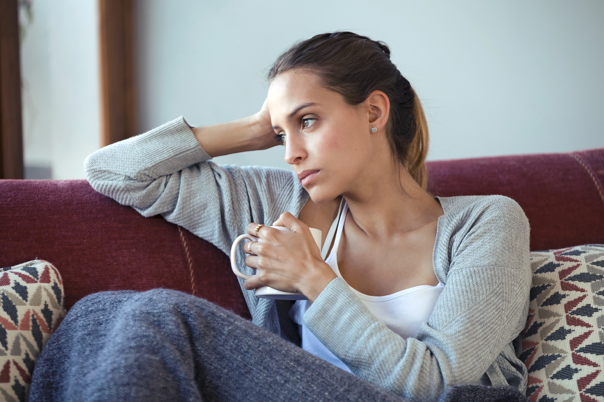 A woman sitting on a red couch holds a mug while looking thoughtfully into the distance. She is wearing a gray cardigan and casual clothes, with her hair tied back. Cushions with geometric patterns are beside her.