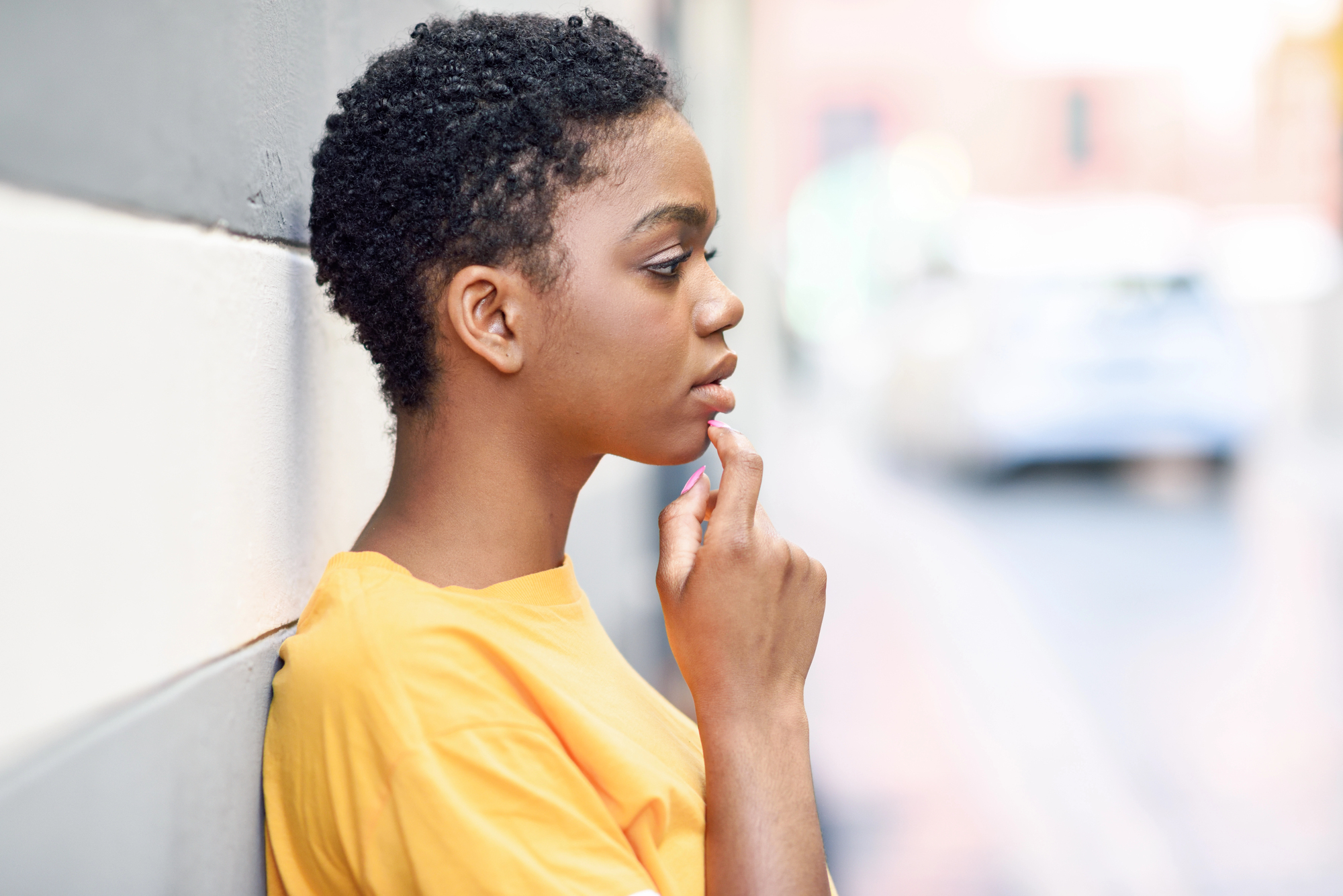 A person with short curly hair and wearing a yellow shirt is leaning against a wall, looking thoughtful. The background is a blurred street scene.