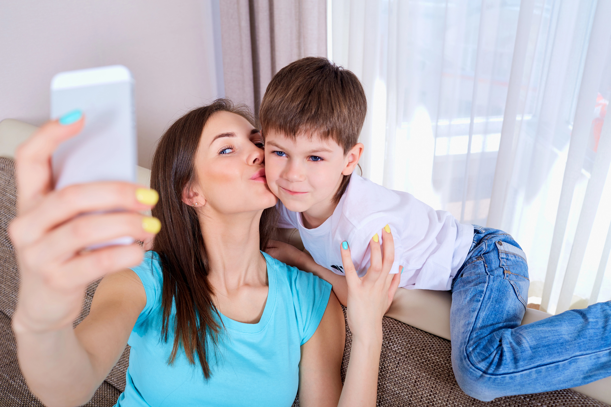 A woman is taking a selfie while giving a young boy a kiss on the cheek. They are sitting on a couch near a window with sheer curtains. Both people are smiling slightly, and the woman is holding the phone with a colorful manicure.