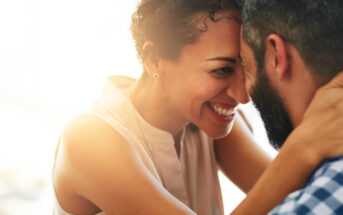 A smiling woman embraces a bearded man, both leaning their foreheads together in a close, affectionate gesture. The background is softly lit, creating a warm and intimate atmosphere.