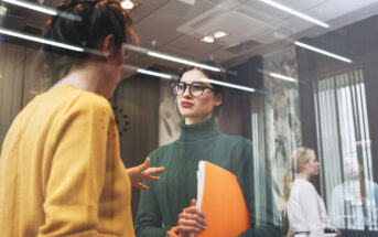 Two women are talking in an office setting. One wears a yellow sweater and gestures, while the other, in a green turtleneck and glasses, holds orange folders. Two more people are seen through a glass partition in the background.