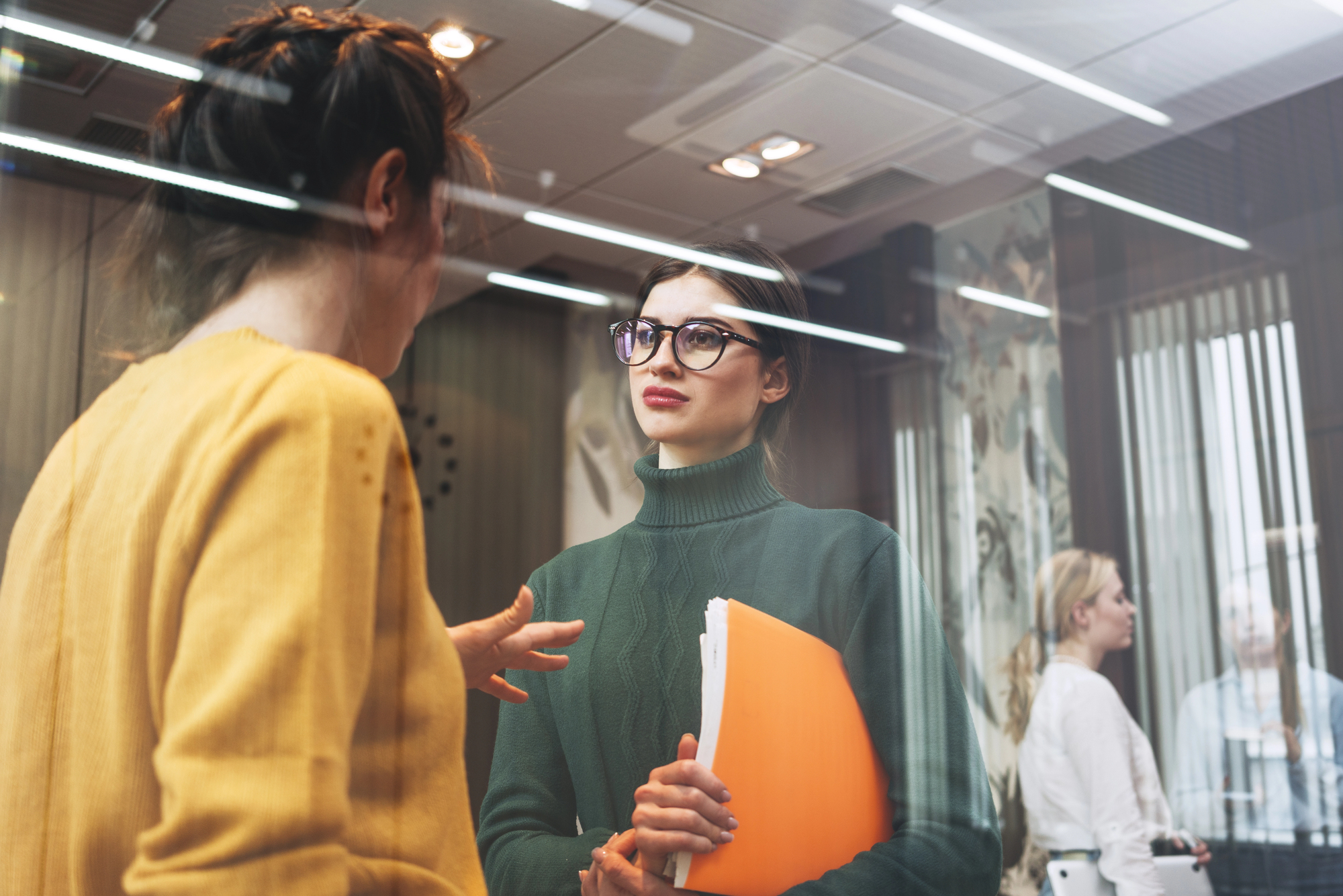 Two women are talking in an office setting. One wears a yellow sweater and gestures, while the other, in a green turtleneck and glasses, holds orange folders. Two more people are seen through a glass partition in the background.