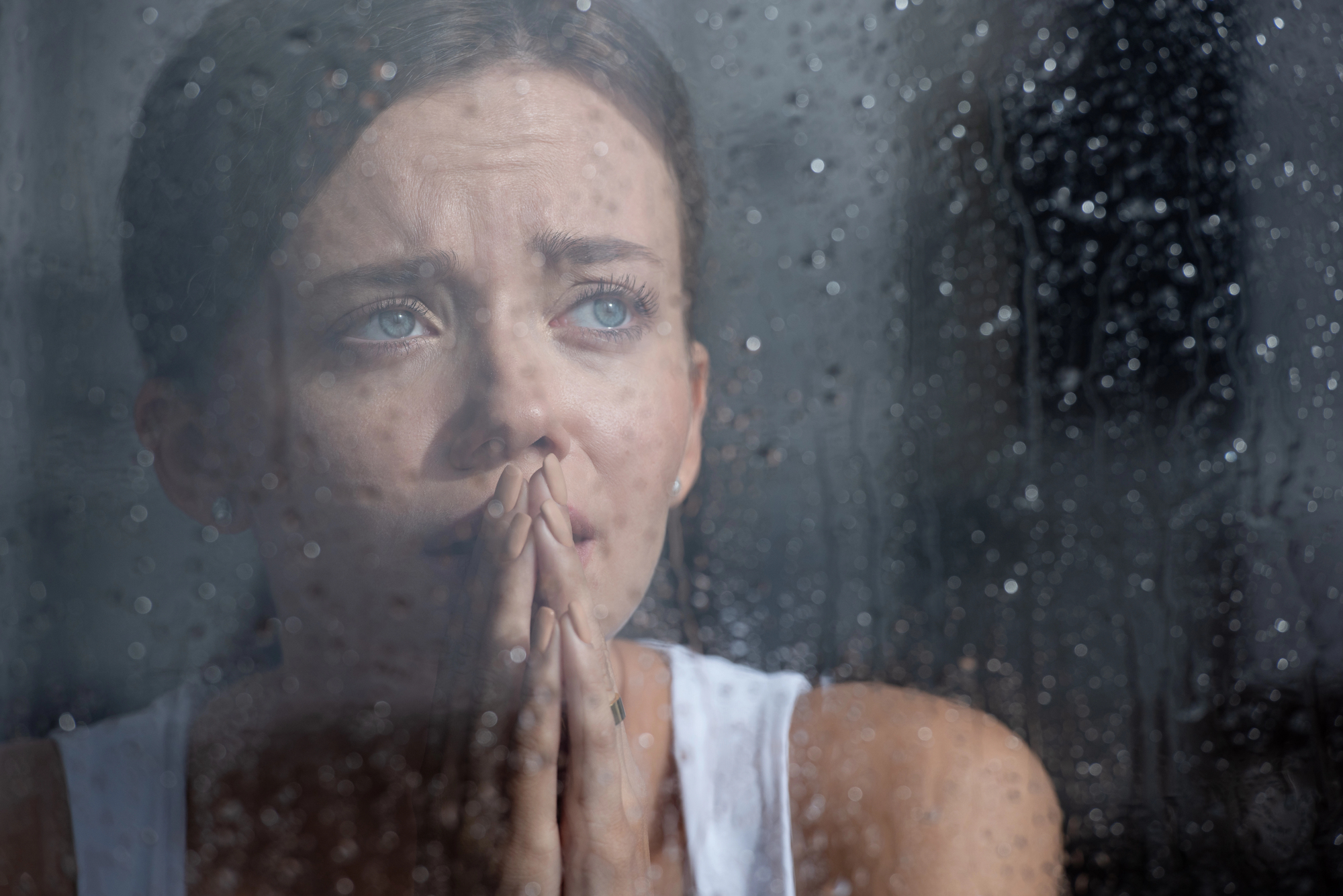 A woman with a contemplative expression is looking through a rain-covered window. Her hands are clasped near her face as she gazes outside, suggesting a sense of longing or reflection. She is wearing a white tank top.