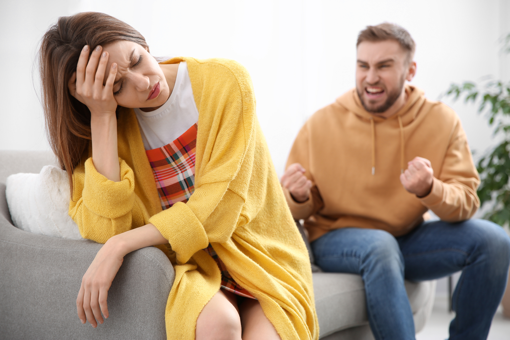 A woman in a yellow cardigan sits on a couch with her head resting on her hand, looking distressed. Behind her, a man in a brown hoodie sits with clenched fists, appearing angry. The background is a bright room with green plants.