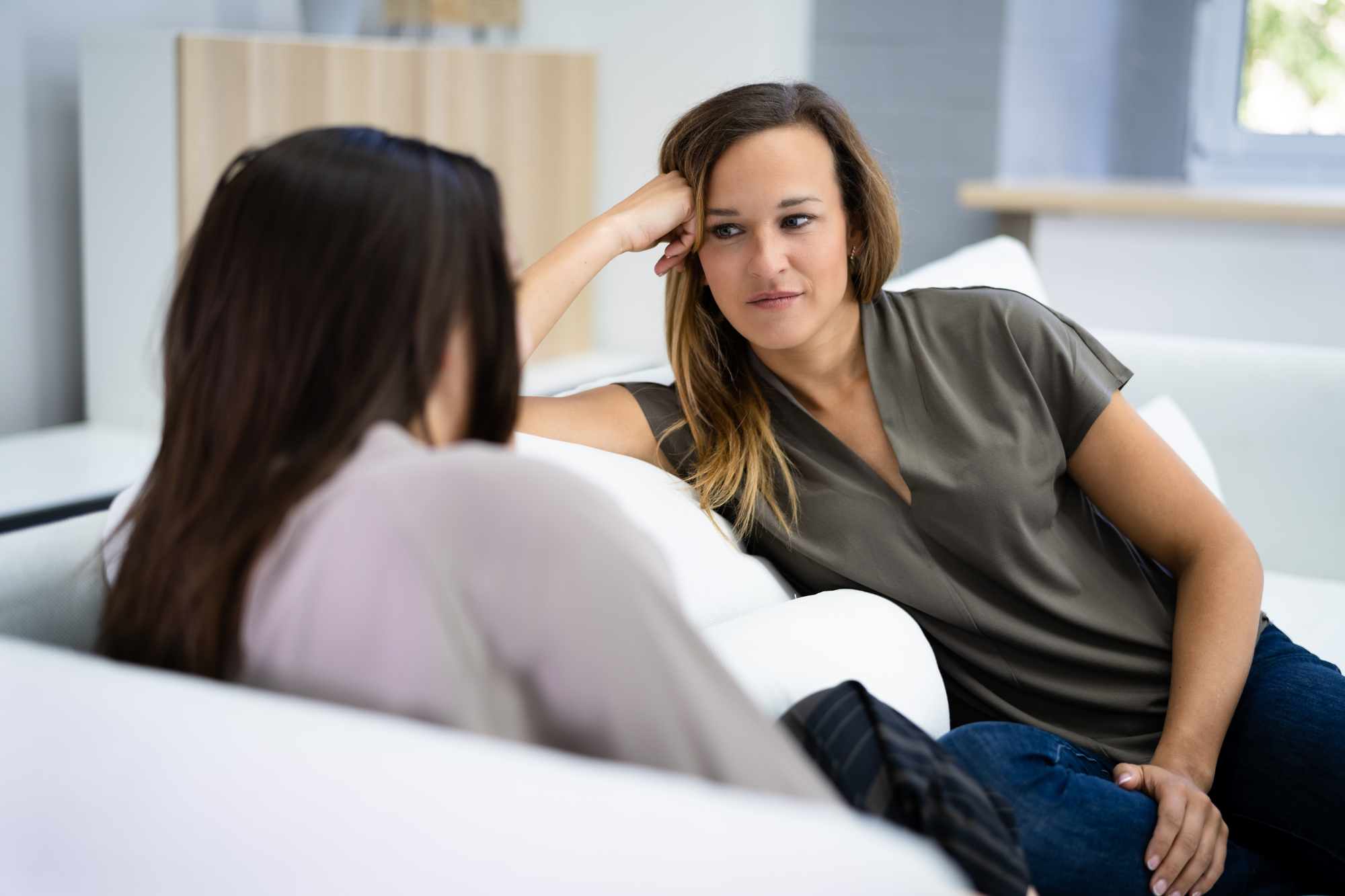 Two women are seated on a white sofa in a well-lit room. One woman with long hair is attentively listening to the other, leaning on her arm. They both appear engaged in conversation, with neutral expressions. A wooden shelf is blurred in the background.