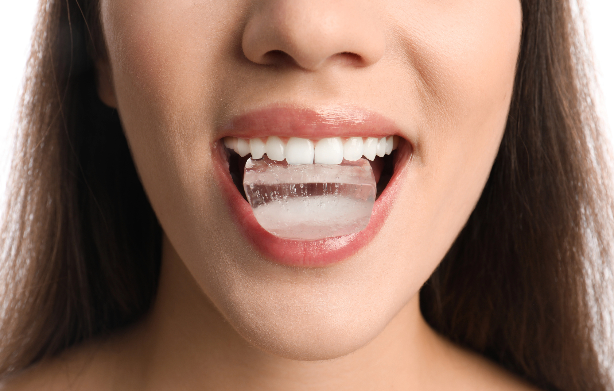 A close-up of a person holding a small ice cube with their teeth, visible through slightly parted lips. The image focuses on the mouth and clean, white teeth. The person has long brown hair.