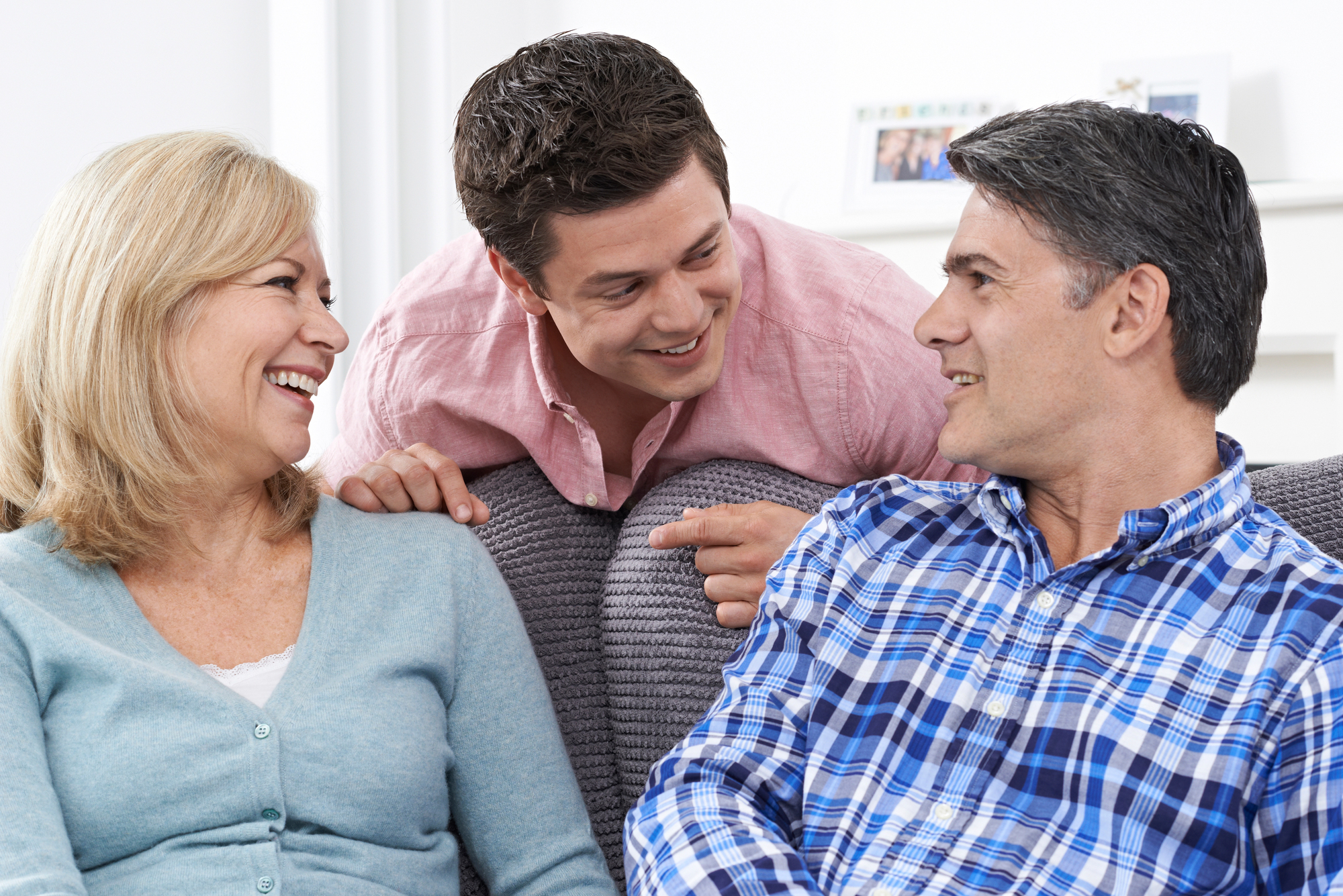 A young man leans over a couch, smiling and talking with an older couple seated on it. The woman on the left wears a light blue sweater, and the man on the right wears a blue plaid shirt. They all appear happy and engaged in conversation.