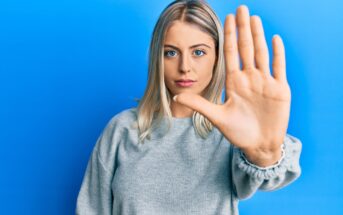 A woman with blonde hair stands against a blue background, extending her hand toward the camera in a stop gesture. She wears a gray sweater and has a neutral expression.