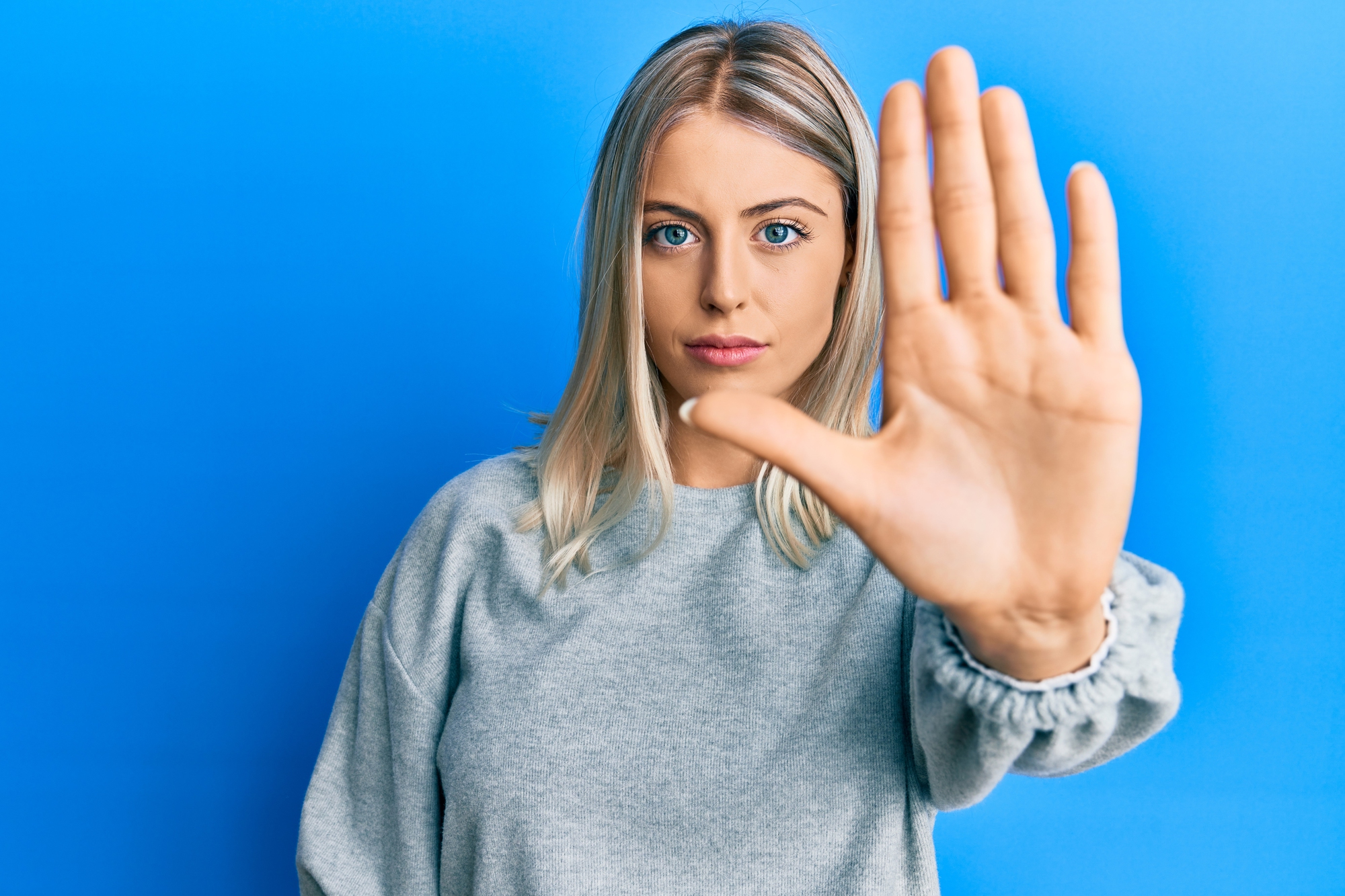 A woman with blonde hair stands against a blue background, extending her hand toward the camera in a stop gesture. She wears a gray sweater and has a neutral expression.