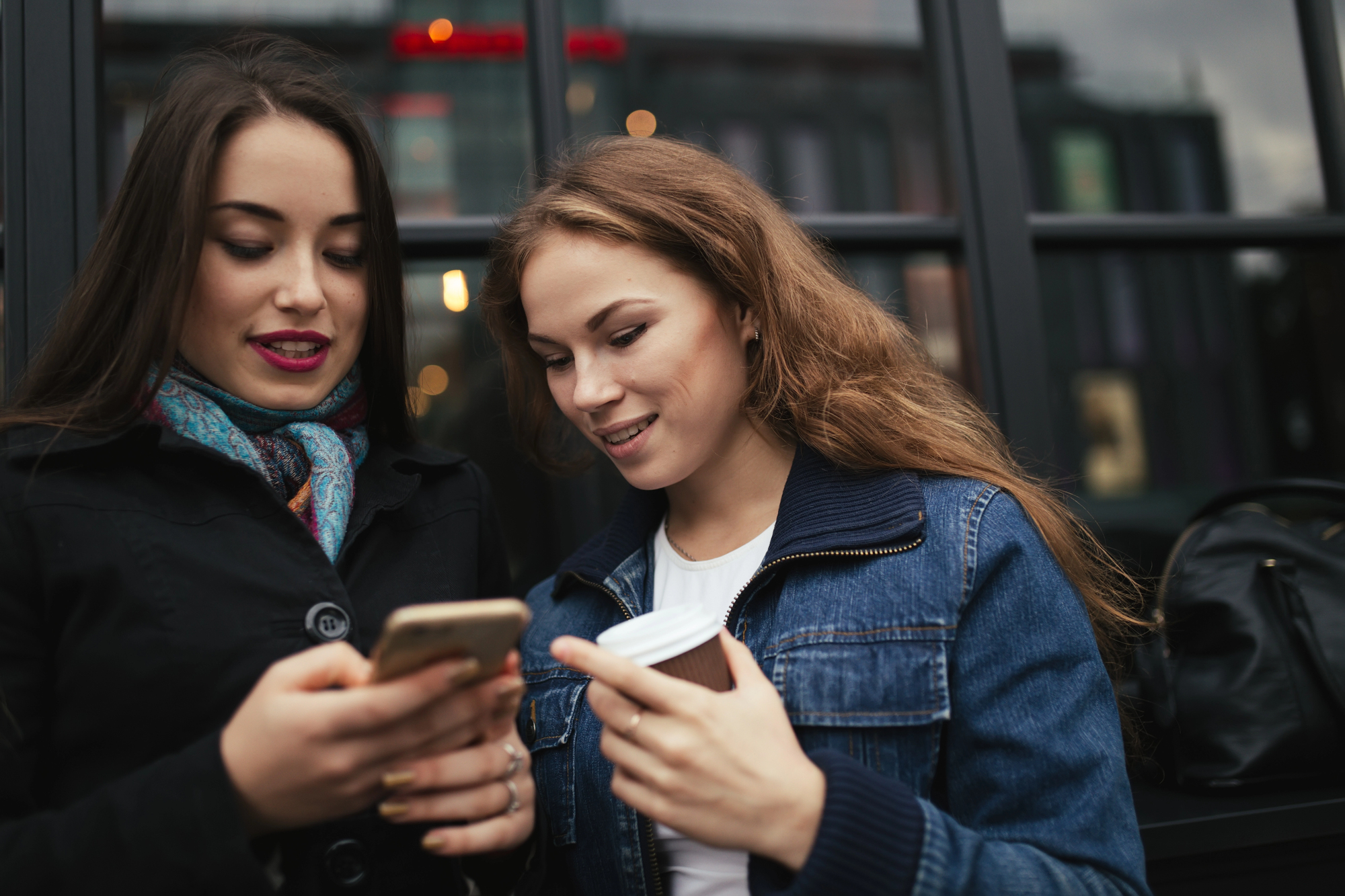 Two women stand outside a building with glass windows. One holds a smartphone and the other holds a coffee cup, both looking at the screen and smiling. They are dressed in casual jackets, and a backpack is visible in the background.