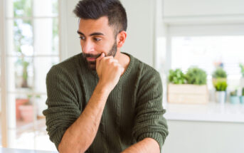 A man with a beard and dark hair, wearing a green sweater, sits pensively at a white table, resting his chin on his hand. The background features potted plants and soft natural light from a nearby window.