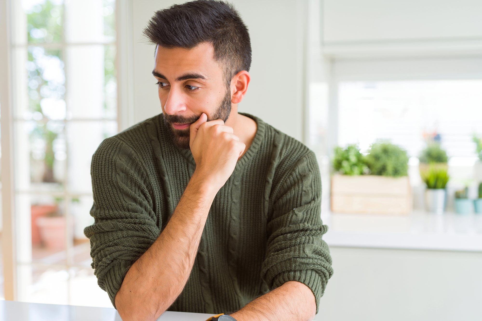 A man with a beard and dark hair, wearing a green sweater, sits pensively at a white table, resting his chin on his hand. The background features potted plants and soft natural light from a nearby window.