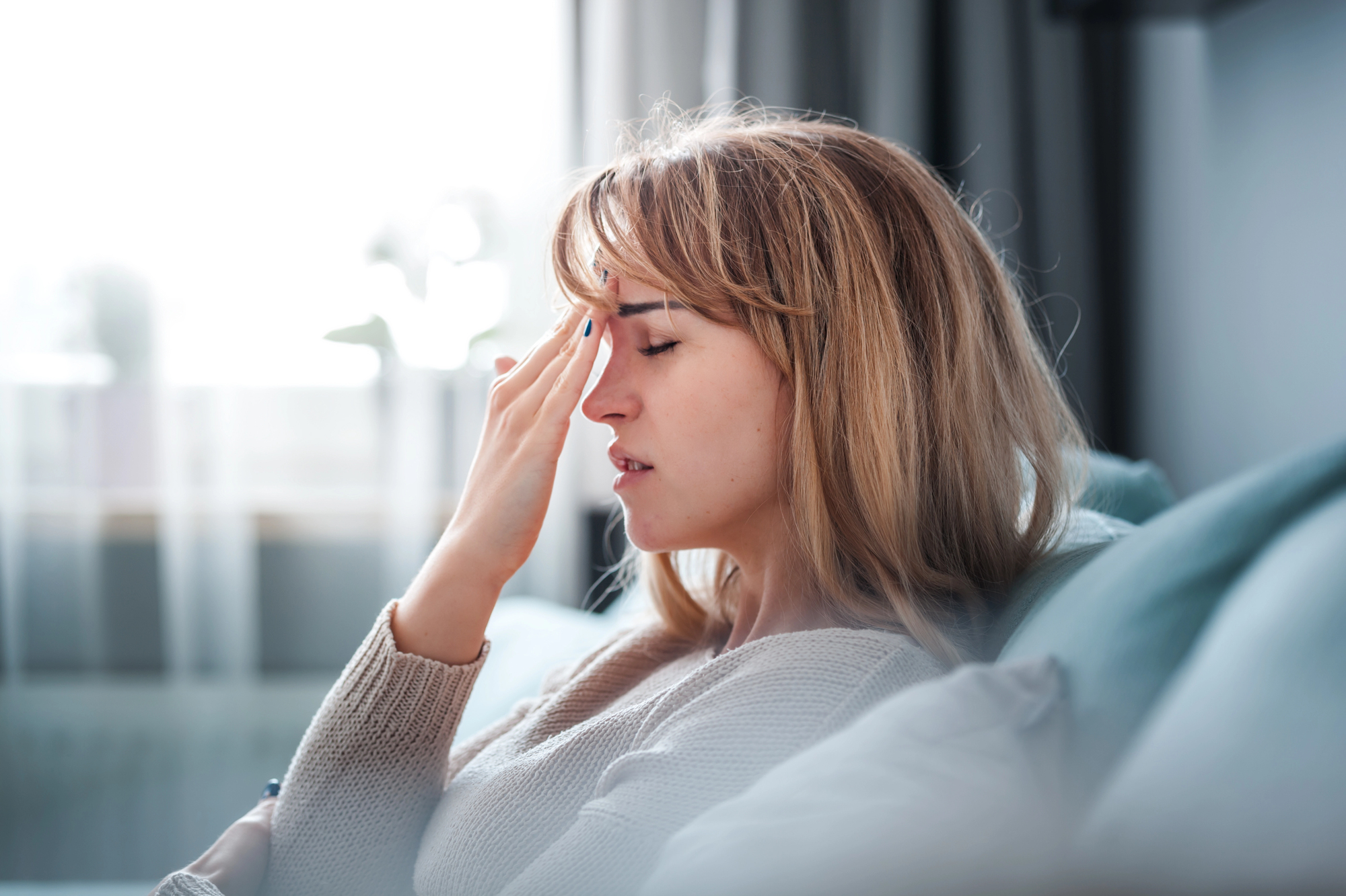 A woman with long blonde hair sits on a couch, eyes closed, gently massaging her temple with her fingers. She appears to be experiencing a headache or stress. The background is softly blurred, suggesting a bright, calm room.