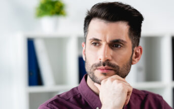 A man with dark hair and a beard wearing a burgundy shirt looks thoughtful, resting his chin on his hand. The background features a bookshelf with folders and a small plant on top.