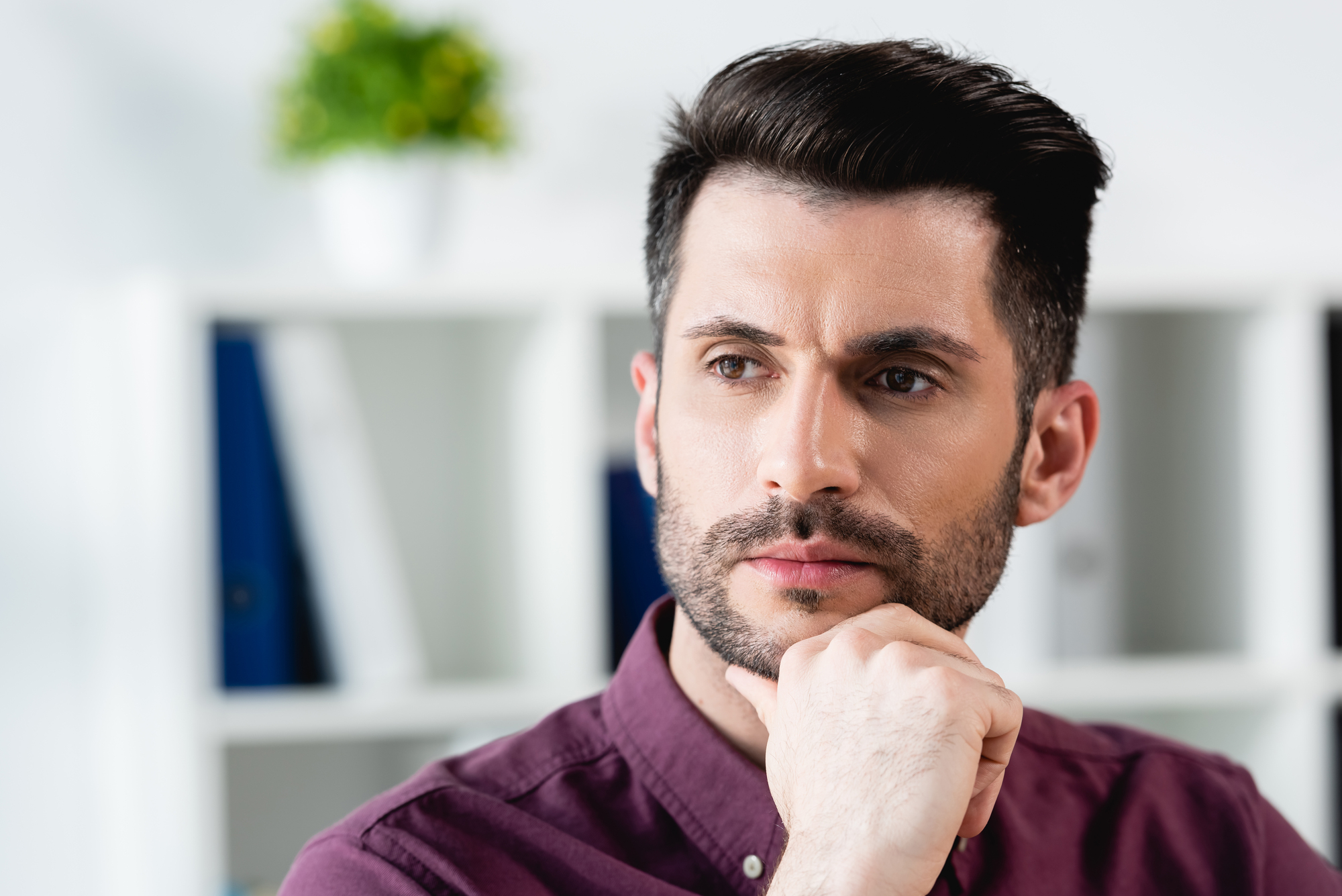 A man with dark hair and a beard wearing a burgundy shirt looks thoughtful, resting his chin on his hand. The background features a bookshelf with folders and a small plant on top.