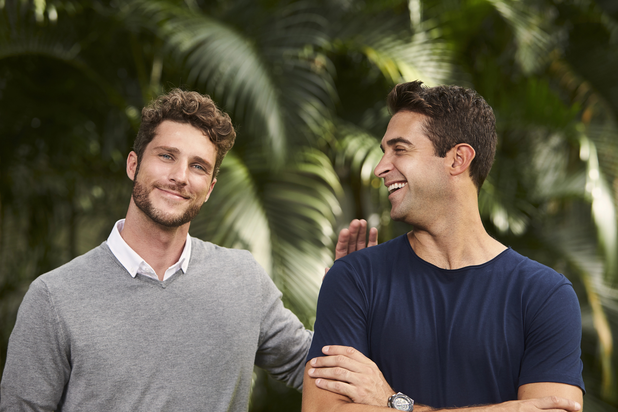 Two men smiling outdoors in front of lush green foliage. One man with curly hair is wearing a gray sweater, while the other has short hair and is dressed in a navy blue t-shirt, appearing to share a light-hearted moment.