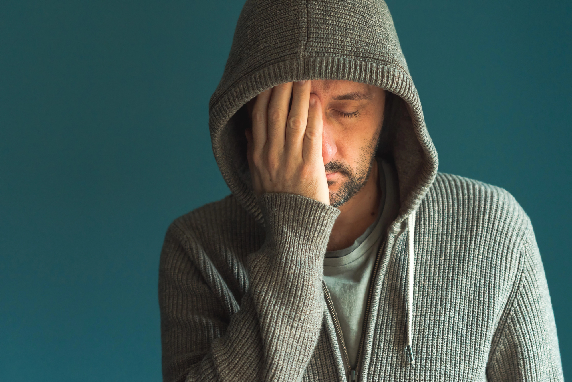 A man wearing a gray hoodie stands against a blue background with his eyes closed and one hand covering half of his face, conveying a sense of exhaustion or frustration.