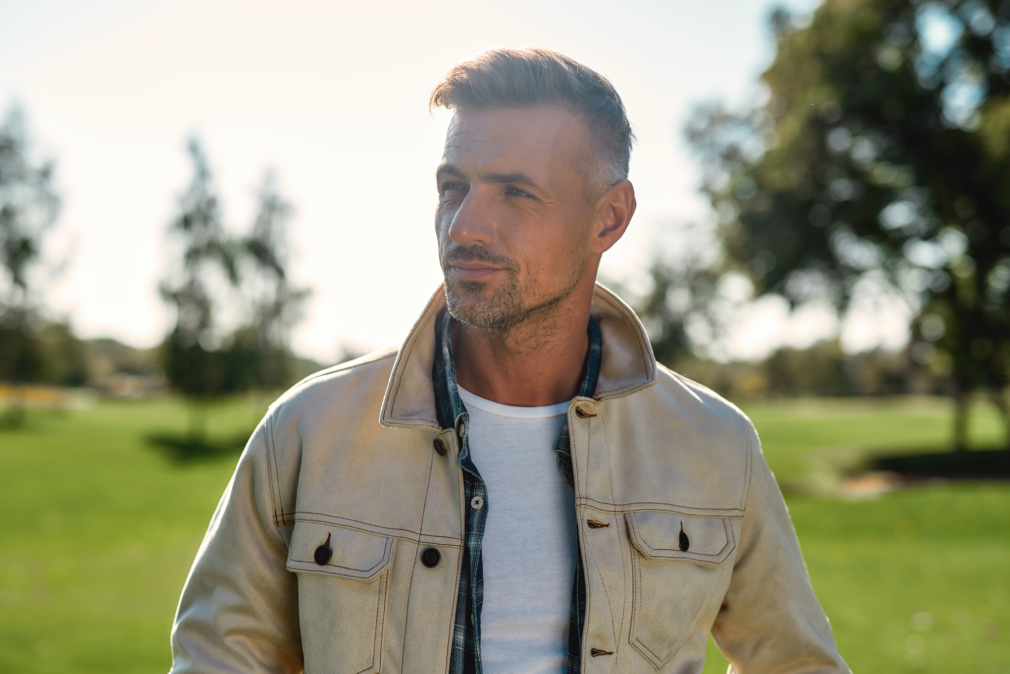 A man with short hair stands outdoors in a sunny park. He is wearing a light-colored jacket over a white shirt. Blurred trees and grass are in the background. The lighting is bright, suggesting a clear day.