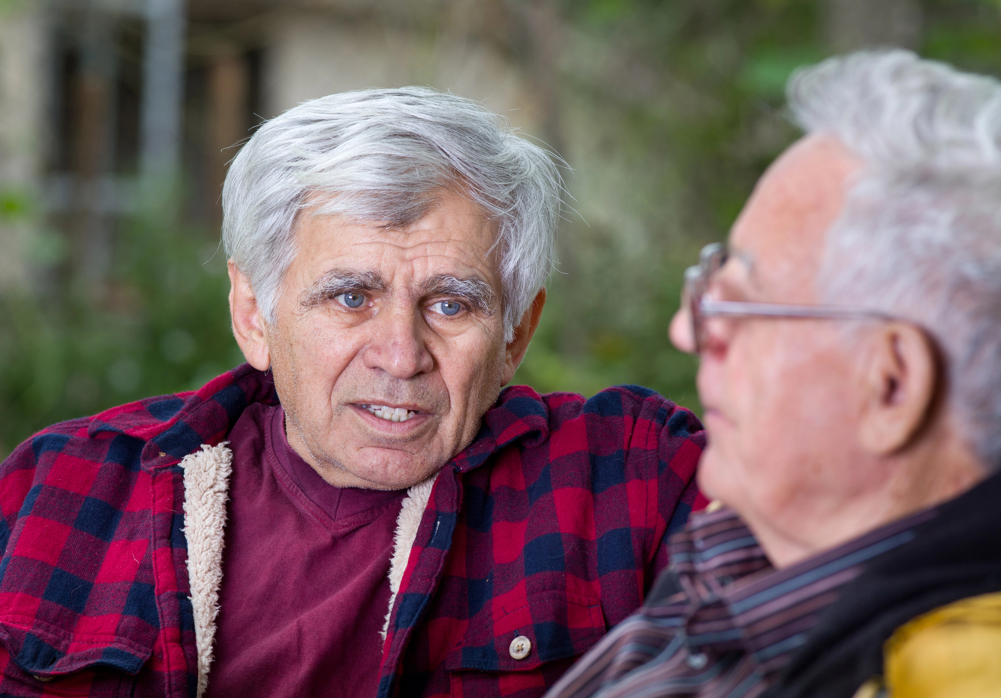 Two elderly men are sitting outdoors, engaged in conversation. One wears a red and black plaid jacket, while the other sports a striped shirt and glasses. The background is blurred with greenery, suggesting a park or garden setting.