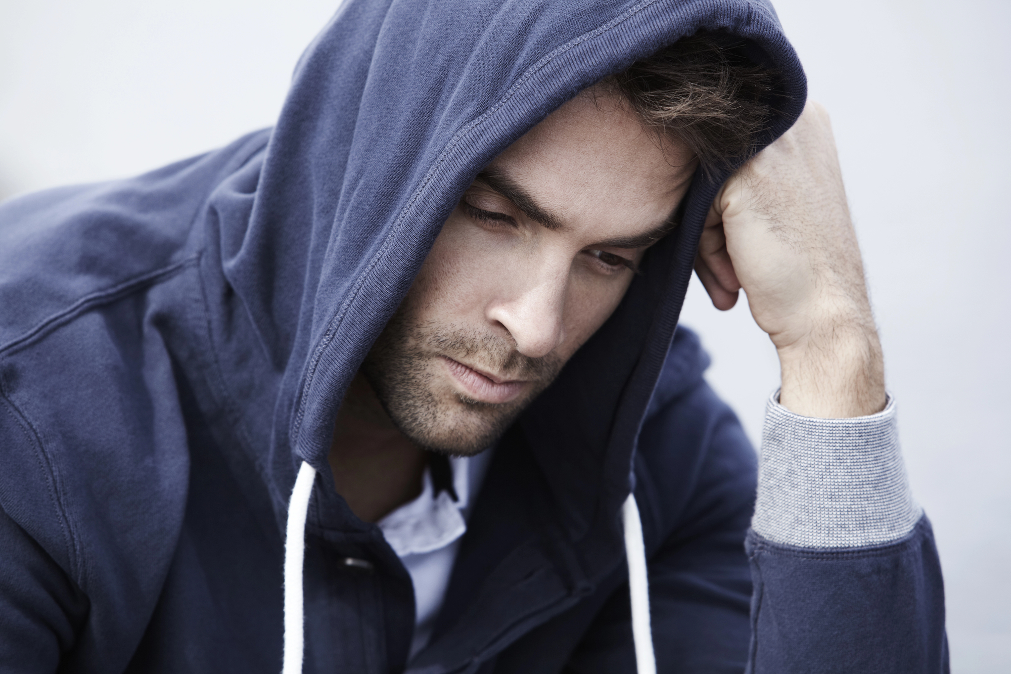 A person wearing a dark blue hoodie is sitting with their head resting on one hand, looking down thoughtfully or pensively. The background is neutral and out of focus.