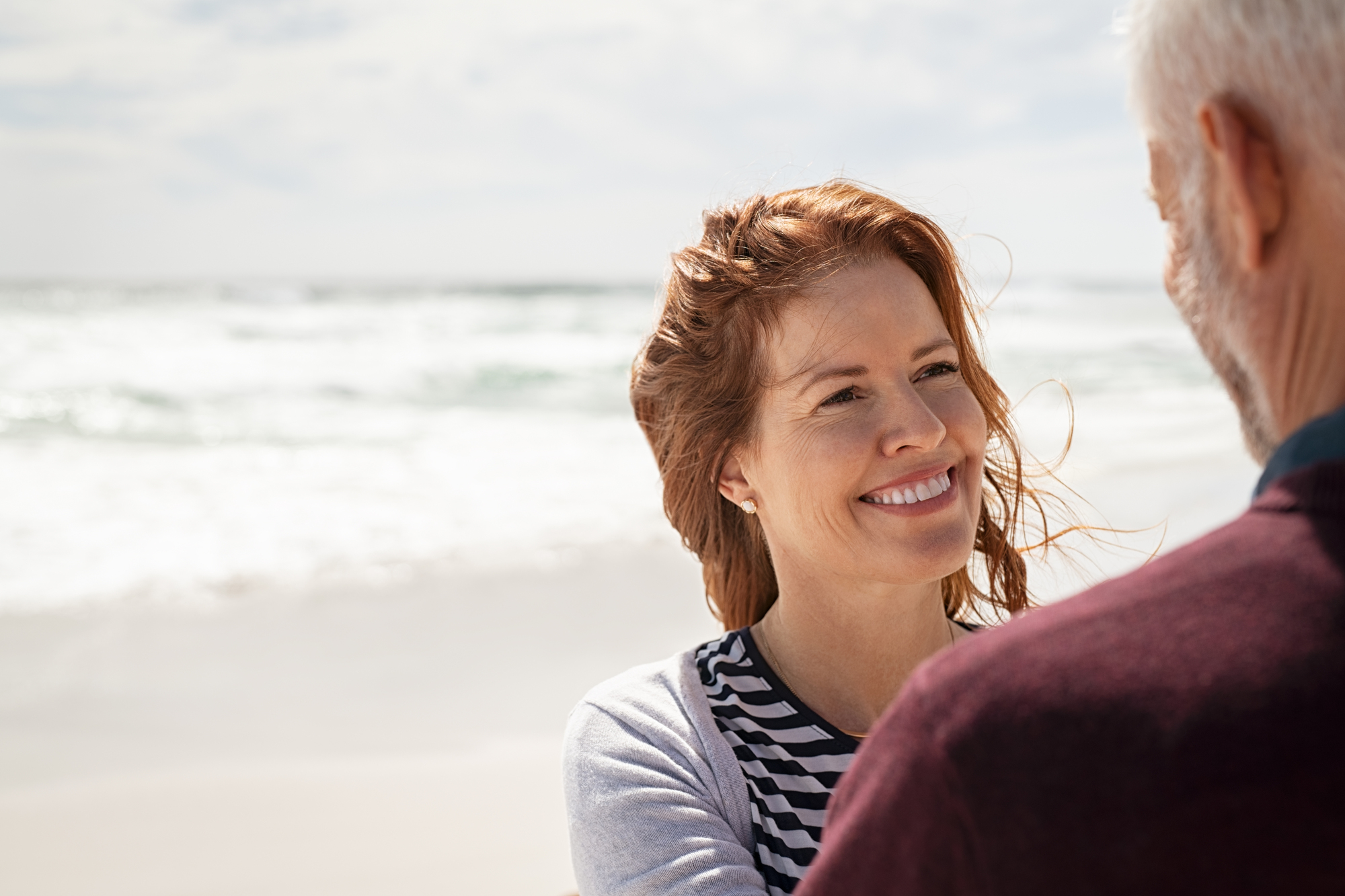 A woman with red hair smiles warmly at a man with gray hair on a sandy beach. The ocean waves are gently rolling in the background under a partly cloudy sky.