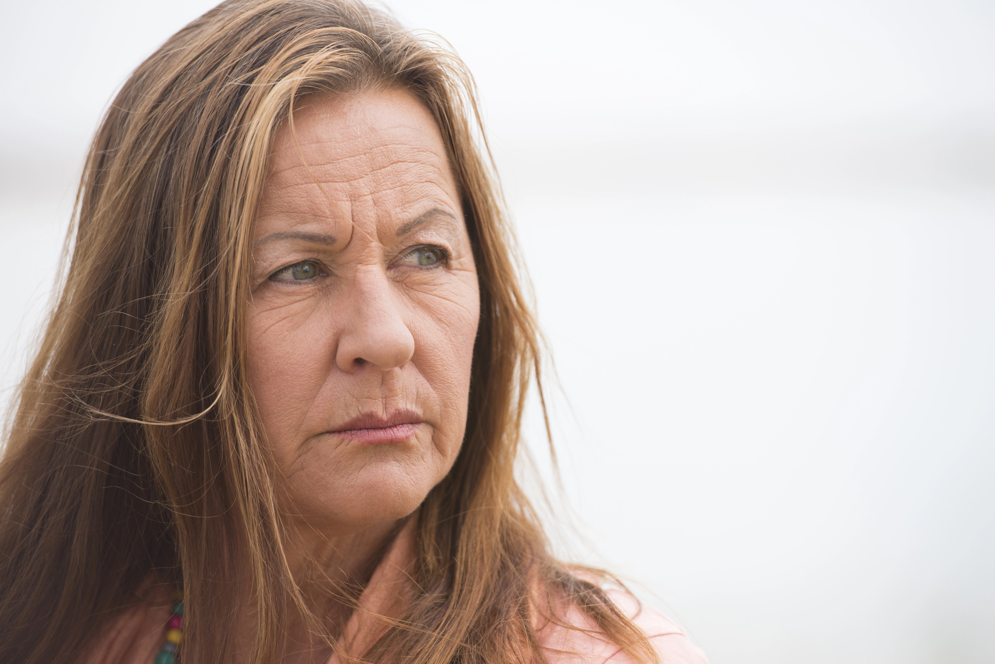 A woman with long brown hair gazes thoughtfully to the side against a light, blurred background. She wears a pink top and necklace, and has a contemplative expression.