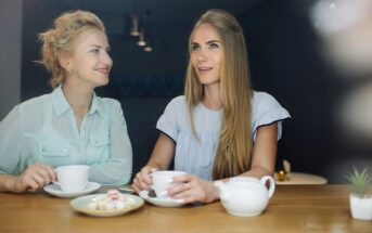 Two women sit at a table in a cafe, smiling and holding cups. One has long blonde hair; the other has a loose bun. A teapot and a plate of small cakes are on the table. The background is softly lit, creating a warm atmosphere.