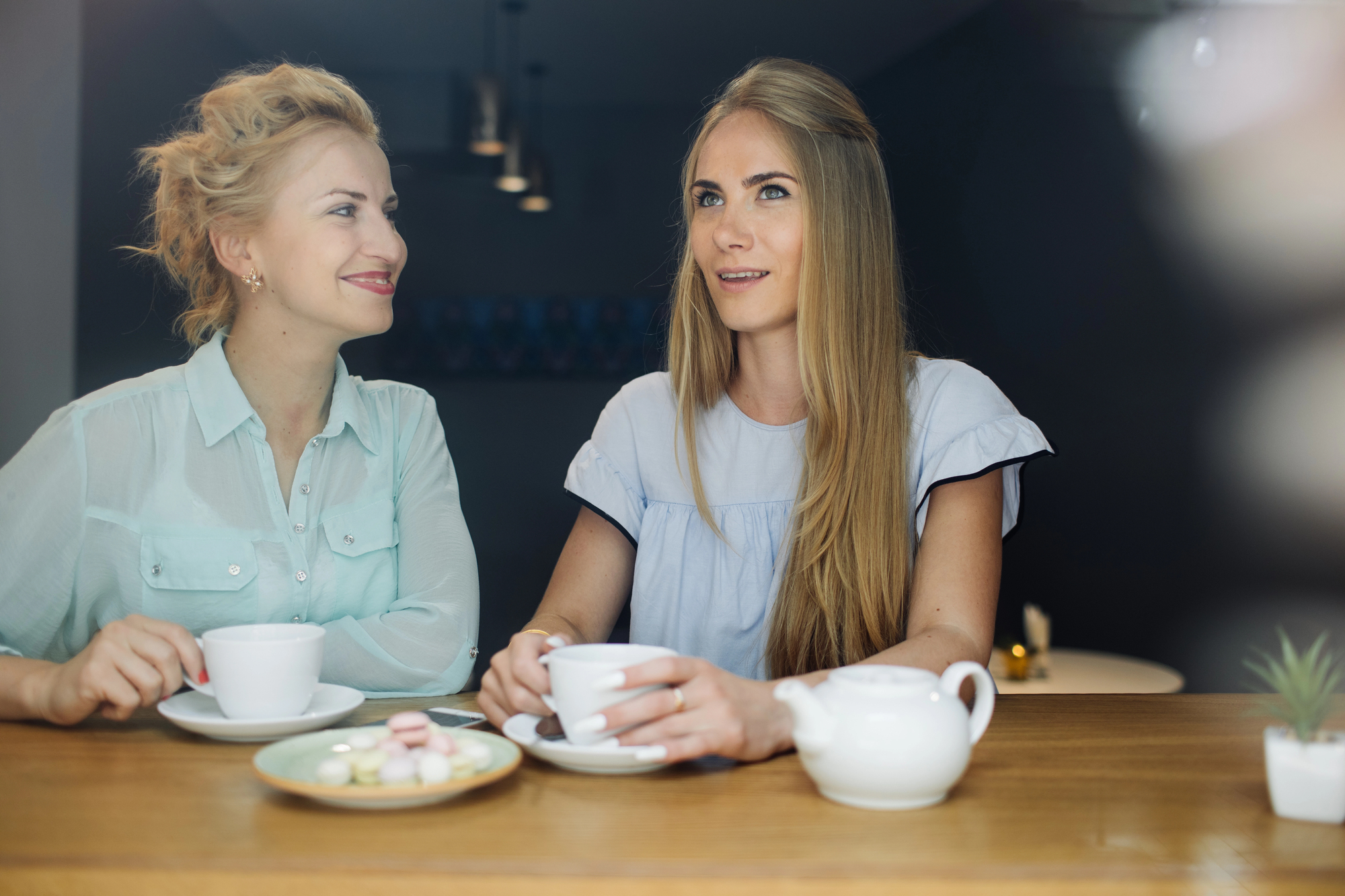 Two women sit at a table in a cafe, smiling and holding cups. One has long blonde hair; the other has a loose bun. A teapot and a plate of small cakes are on the table. The background is softly lit, creating a warm atmosphere.