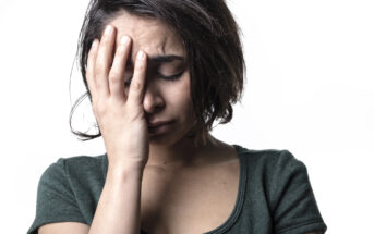 A woman with short dark hair is holding her hand to her forehead, eyes closed, and appears upset or stressed. She is wearing a dark green top against a plain white background.