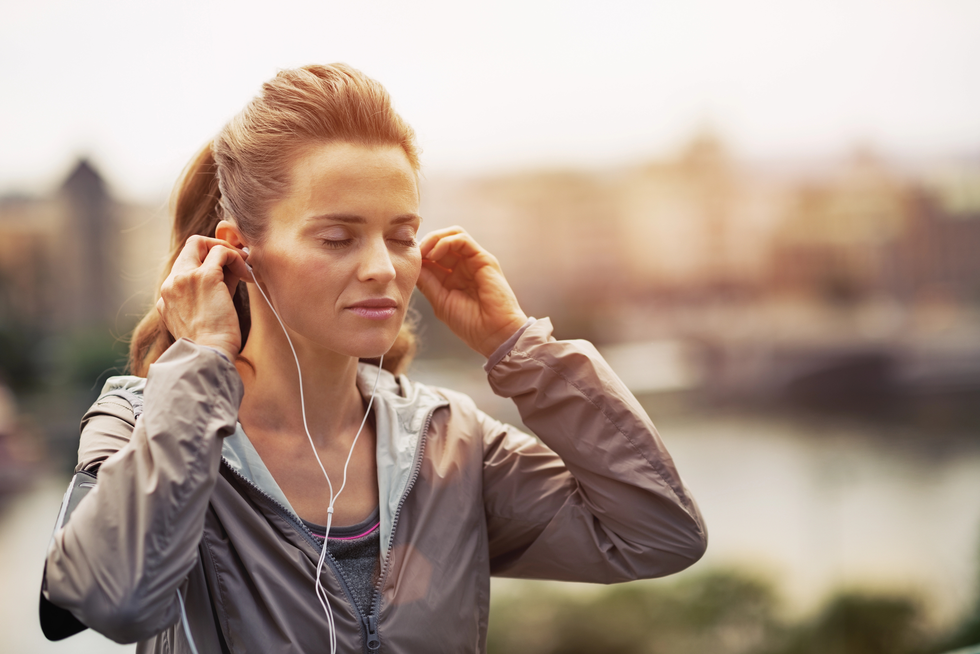 A woman wearing a gray jacket is outdoors, placing earphones in her ears. She has her eyes closed and appears serene, with a blurred cityscape in the background.