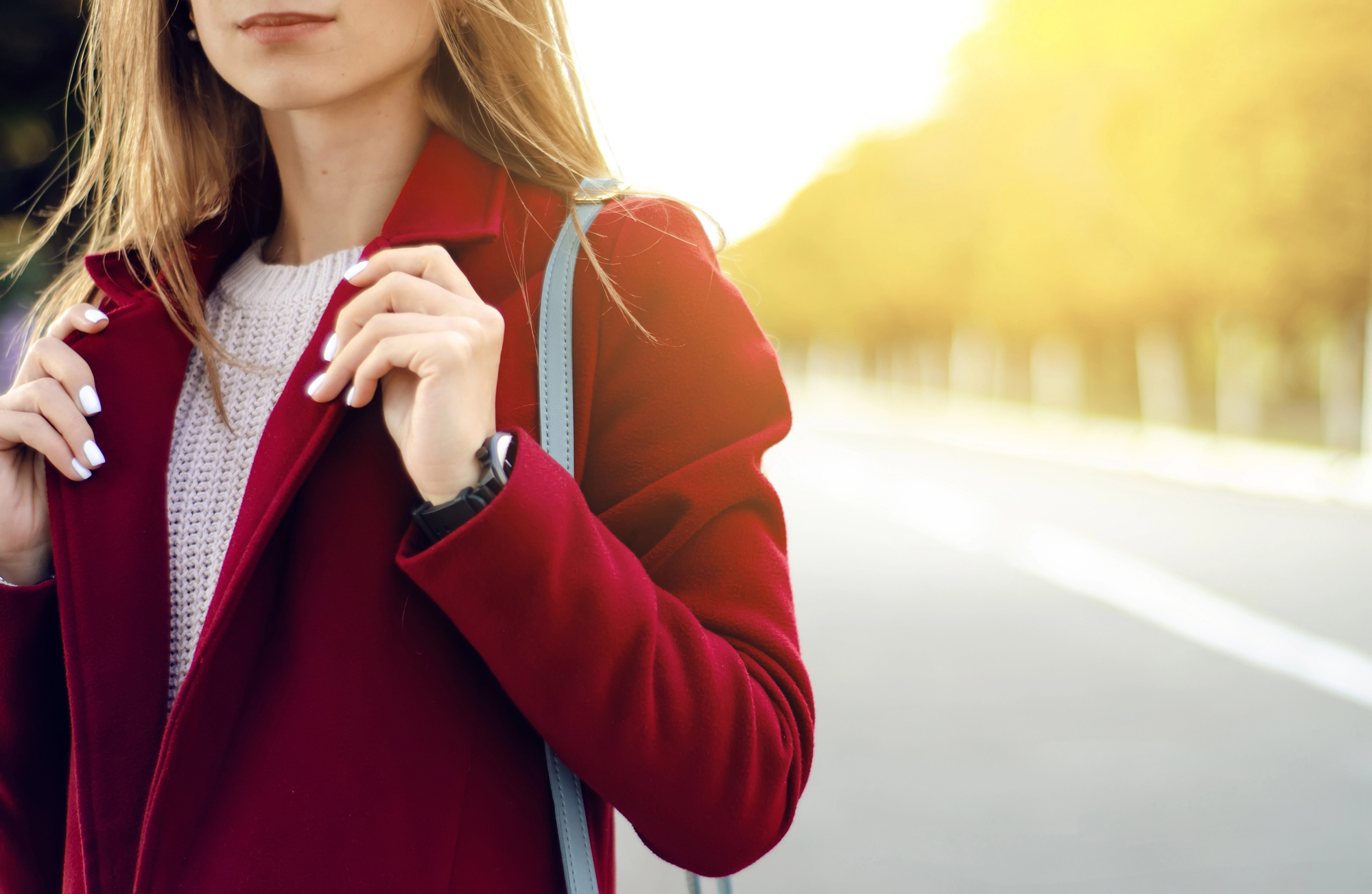 A woman in a red coat stands outdoors on a sunny day, adjusting her collar. She has long hair and is wearing a white sweater underneath. The background is a blurry tree-lined street bathed in golden light.