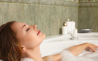 A woman relaxes in a bubble bath, surrounded by lit candles. She has her eyes closed, leaning back against the bathtub. The bathroom walls are tiled with a green pattern.