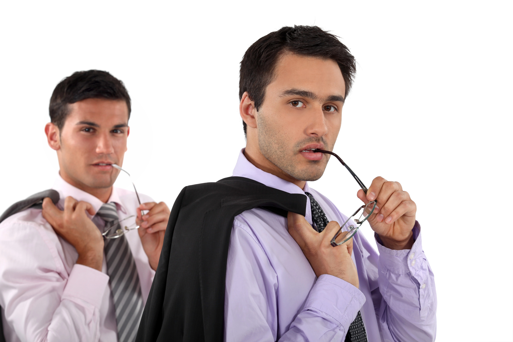 Two men in dress shirts and ties stand confidently against a white background. Each holds a jacket draped over one shoulder. The man in the foreground bites the arm of his glasses, while the other looks ahead, holding his glasses near his face.