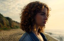 A person with curly hair gazes into the distance while standing on a beach. They are wearing a denim jacket, and the ocean and cliffside are visible in the background, under a partly cloudy sky.