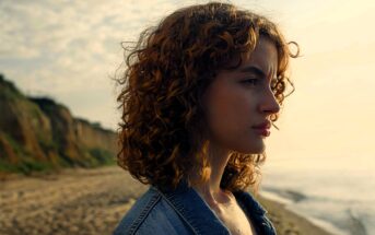 A person with curly hair gazes into the distance while standing on a beach. They are wearing a denim jacket, and the ocean and cliffside are visible in the background, under a partly cloudy sky.