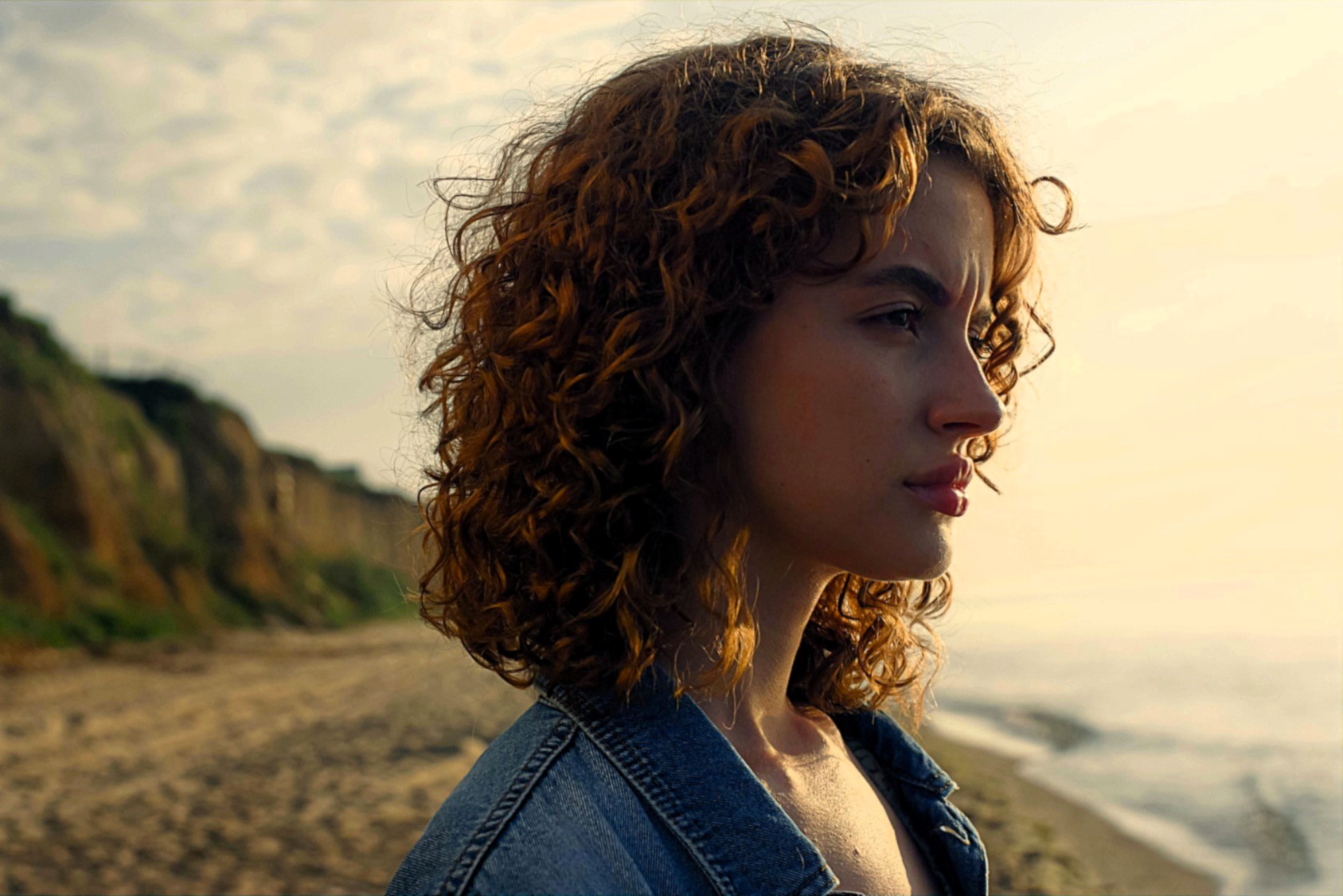 A person with curly hair gazes into the distance while standing on a beach. They are wearing a denim jacket, and the ocean and cliffside are visible in the background, under a partly cloudy sky.