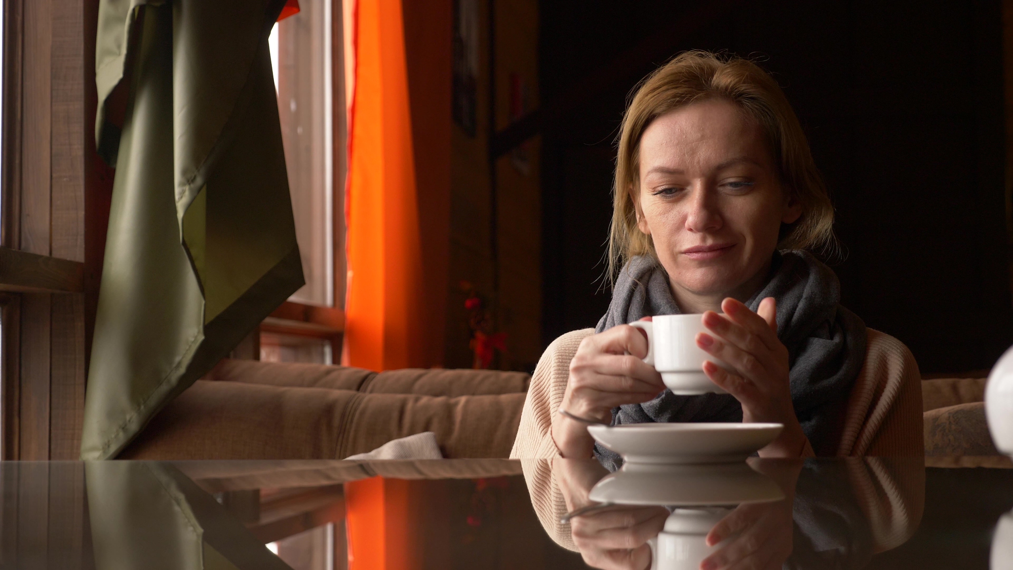 A woman with a scarf sits in a cozy cafe, holding a cup of coffee. She gazes at it thoughtfully, with a warm reflection on the glossy table surface. Sunlight streams through the window, casting soft, natural light on the scene.