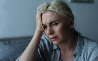 A woman with blonde hair, wearing a gray shirt, appears pensive as she rests her hand on her forehead. She sits indoors with blurred background elements, conveying a sense of contemplation or concern.