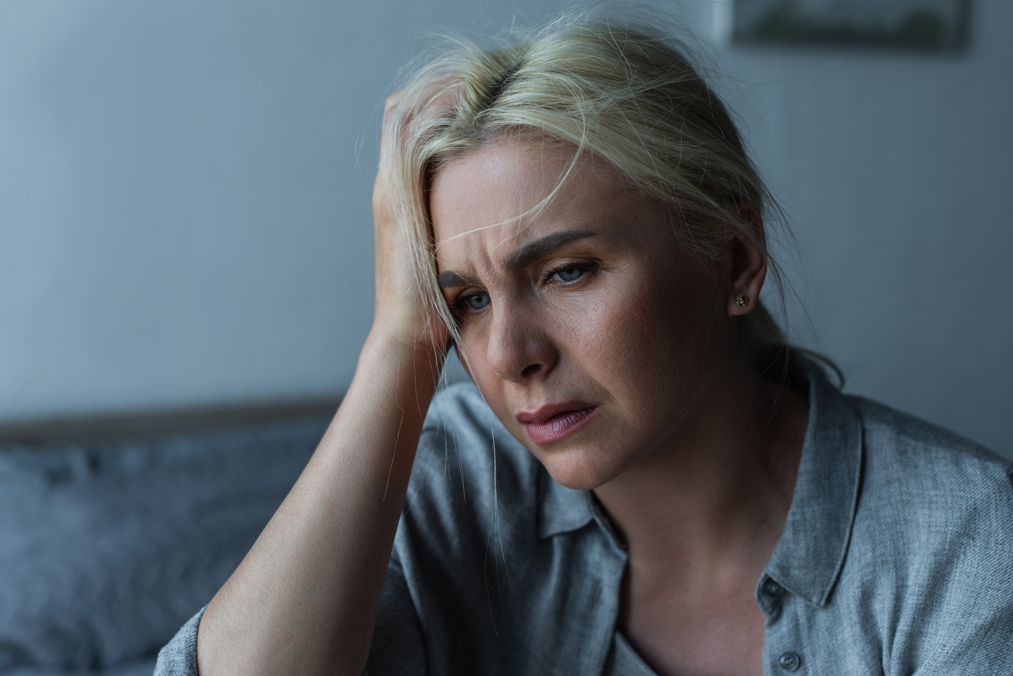 A woman with blonde hair, wearing a gray shirt, appears pensive as she rests her hand on her forehead. She sits indoors with blurred background elements, conveying a sense of contemplation or concern.