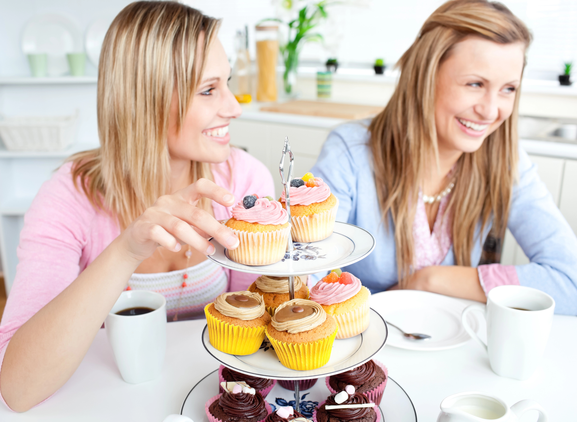 Two women are sitting at a table, smiling and enjoying cupcakes from a three-tiered stand. The cupcakes are decorated with various frostings. Two white coffee cups are also on the table. The background shows a bright, modern kitchen.