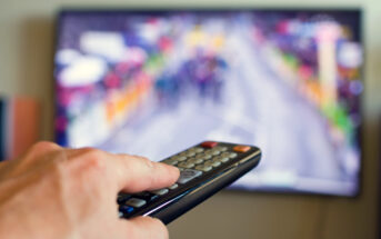 A hand holding a TV remote points towards a television displaying a blurred image of a colorful outdoor scene, possibly a race or parade, with people and banners on the screen.